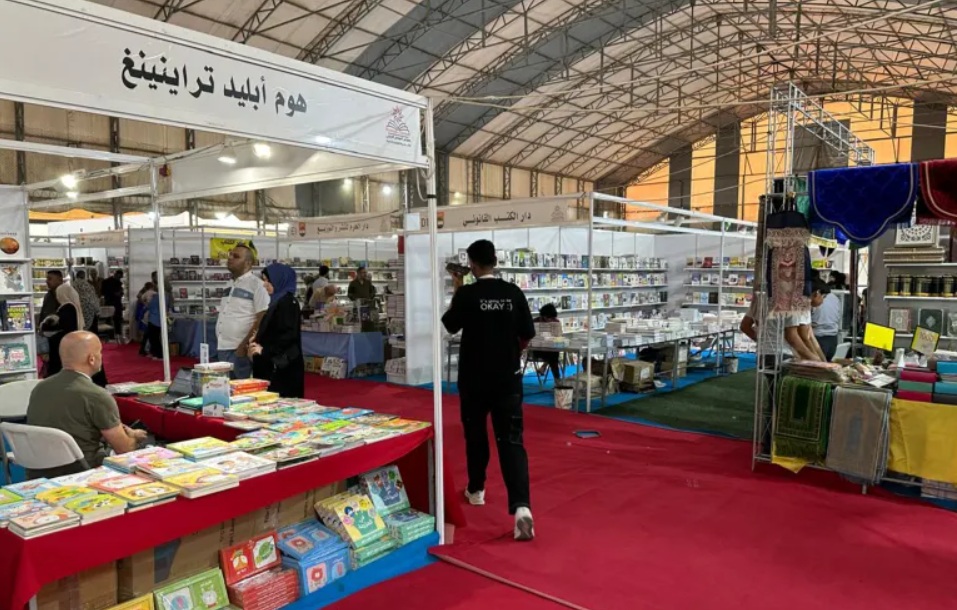 A book fair in Mosul under a large tent, with rows of bookseller stalls, displays of books in Arabic, and visitors browsing the tables.
